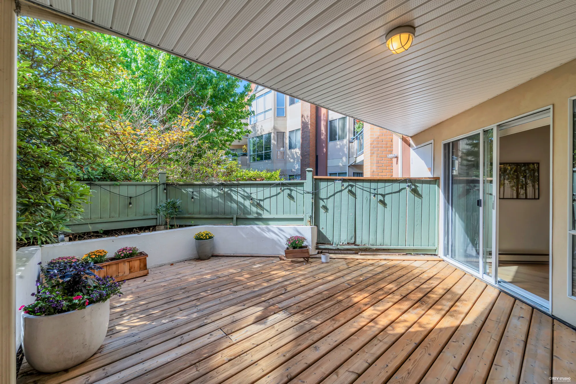 The private deck at 151-8600 Lansdowne Road: wood boards, a covered section, teal railings and the garden beyond
